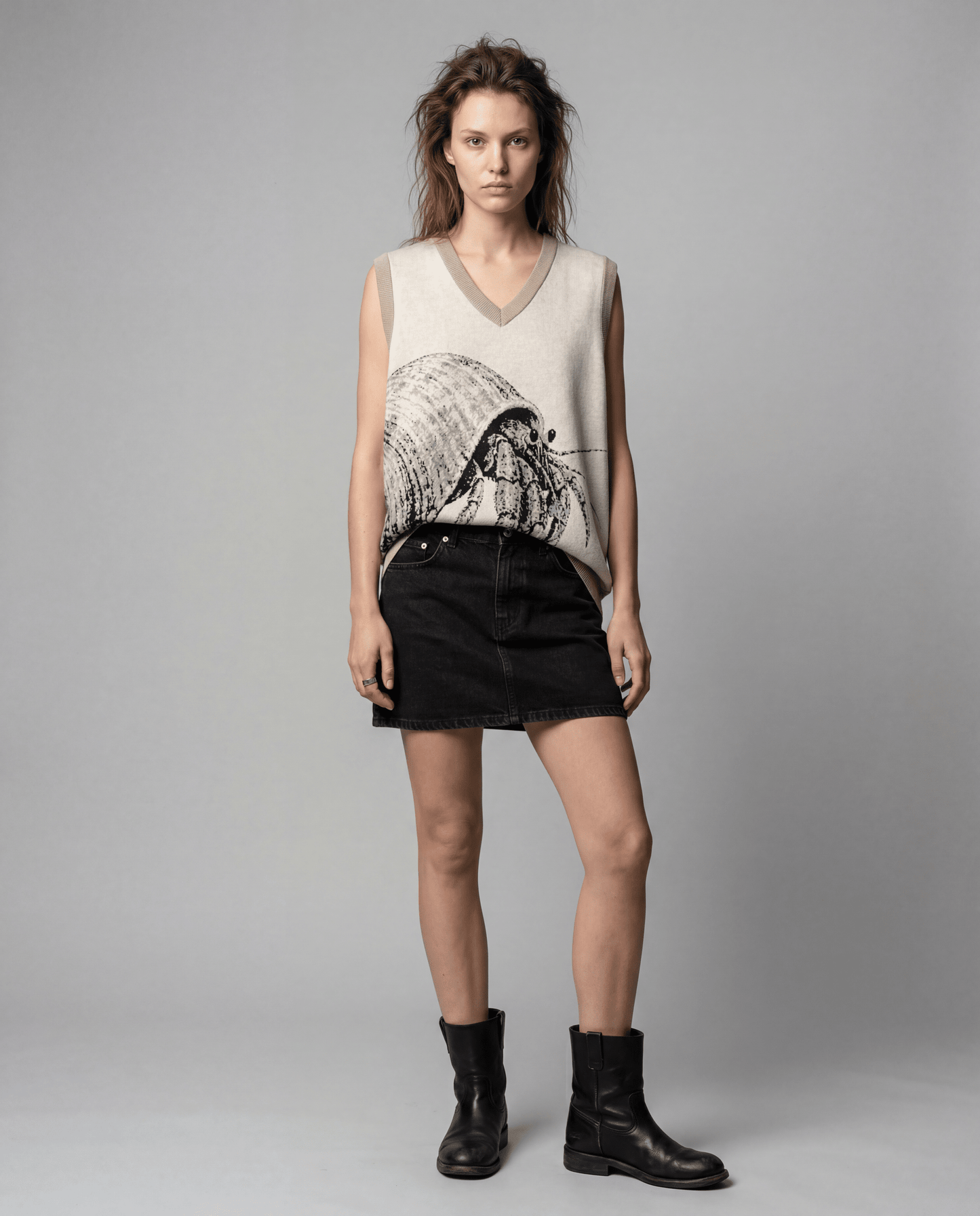 Model wearing light beige sleeveless knit vest with hermit crab design styled with high-waisted black denim mini skirt