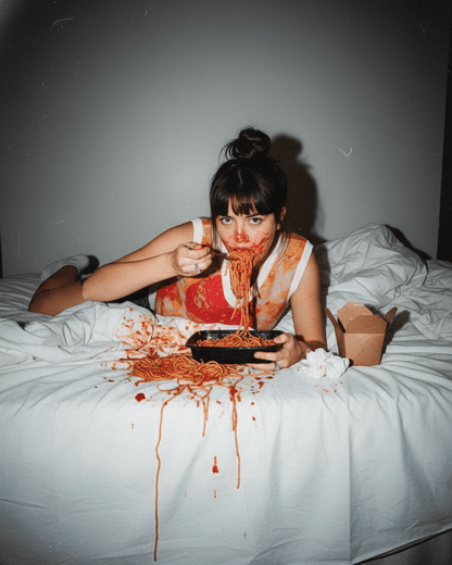 Person with spaghetti on their face and clothes, sitting on a bed.