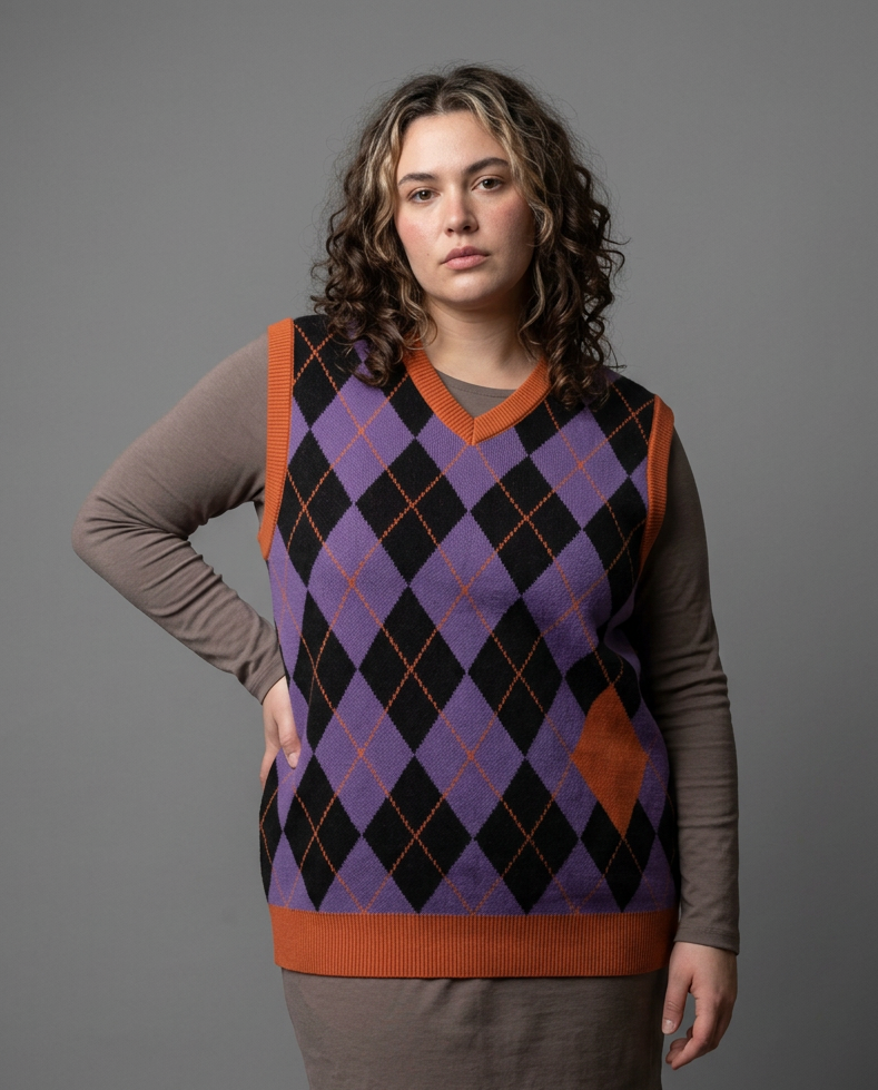 A woman modeling a sleeveless argyle sweater vest over a brown long-sleeve top and matching brown skirt, photographed against a solid gray studio background.